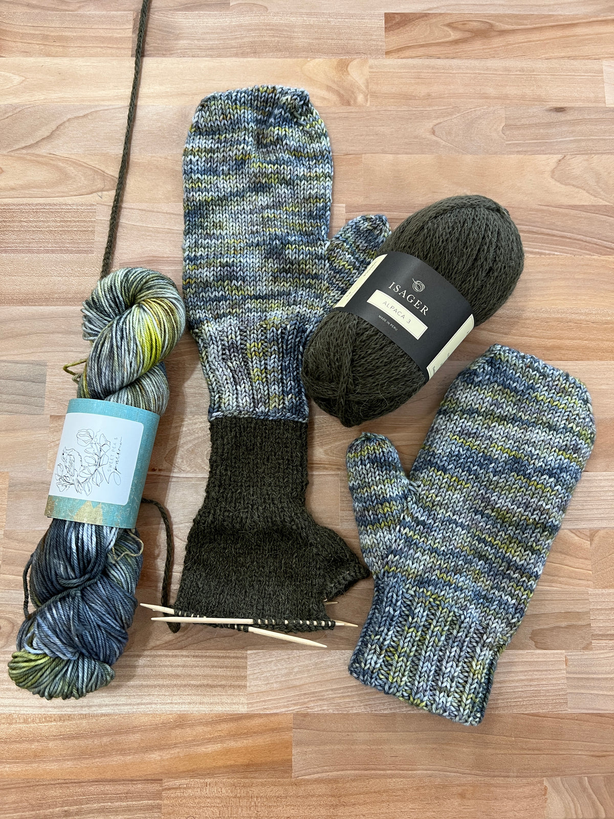 Hand-knitted, lined mittens in a variegated blue, green, and gray yarn, displayed on a light wood floor alongside two skeins of matching yarn and knitting needles.