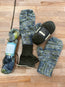 Hand-knitted, lined mittens in a variegated blue, green, and gray yarn, displayed on a light wood floor alongside two skeins of matching yarn and knitting needles.