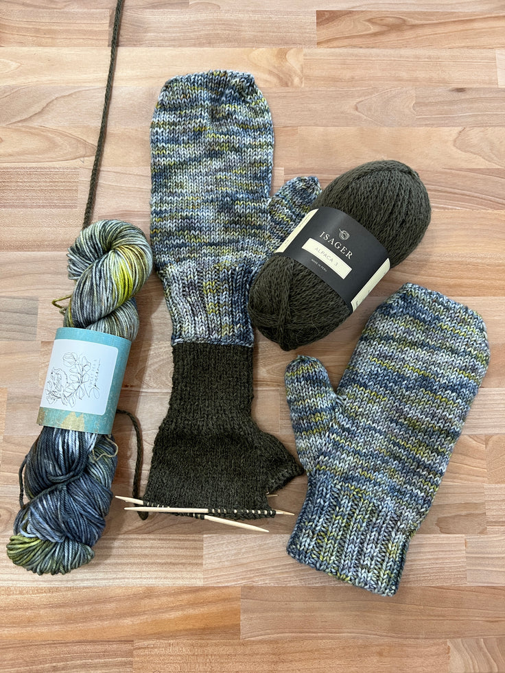 Hand-knitted, lined mittens in a variegated blue, green, and gray yarn, displayed on a light wood floor alongside two skeins of matching yarn and knitting needles.